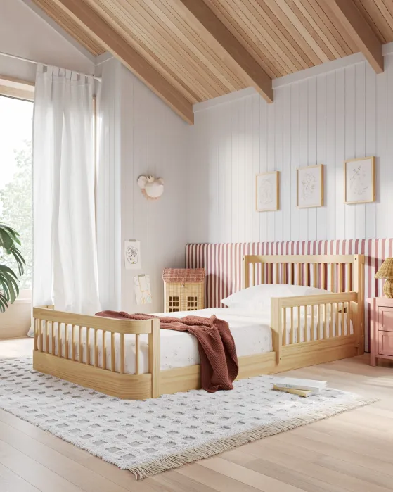 A cozy bedroom with a wooden bed, white bedding, a rust-colored blanket, striped headboard, framed art on the wall, and a large window with sheer curtains.
