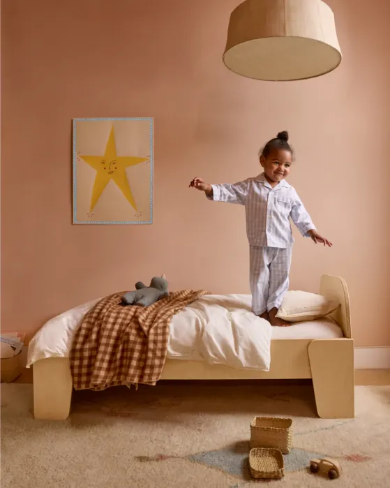 Young child in pajamas stands on a bed in a softly lit room with tan walls, a star poster, toys on the floor, and a blanket draped over the bed.