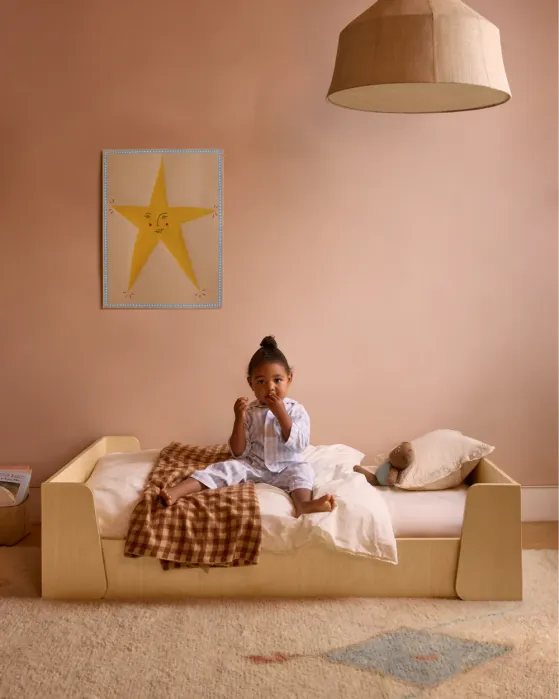A young child in pajamas sits on a wooden bed in a softly lit bedroom with peach walls, a star poster, a pillow, a brown blanket, and a stuffed toy.