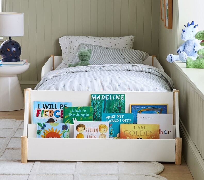 A child's bedroom with a white bed and star-patterned bedding, a bedside lamp, and a low bookshelf holding several children's books at the foot of the bed.