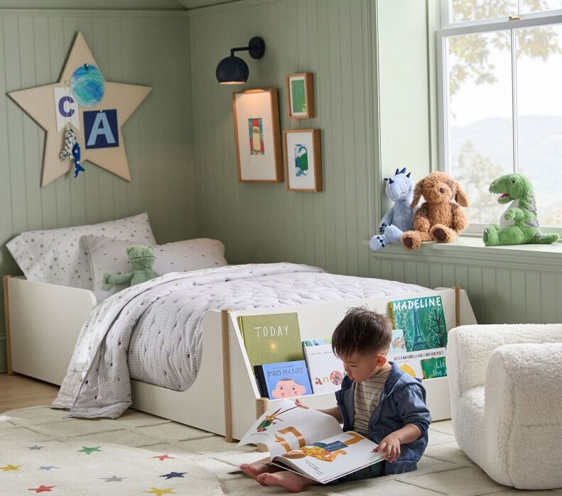 A young child sits on the floor reading a book in a neatly organized, pastel-green bedroom with a bed, plush toys, and a bookshelf filled with children's books.