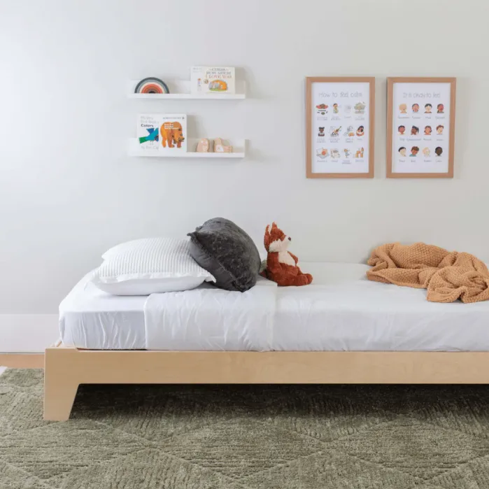 A neatly made bed with white sheets, pillows, and a stuffed fox toy sits against a white wall with two framed posters and shelves holding books and toys above it.