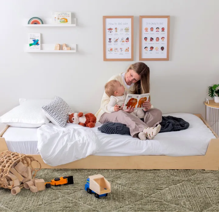 A woman sits on a child's bed reading a book to a toddler. Wooden toys and a stuffed animal are on the bed and floor. Children's posters and shelves decorate the wall.