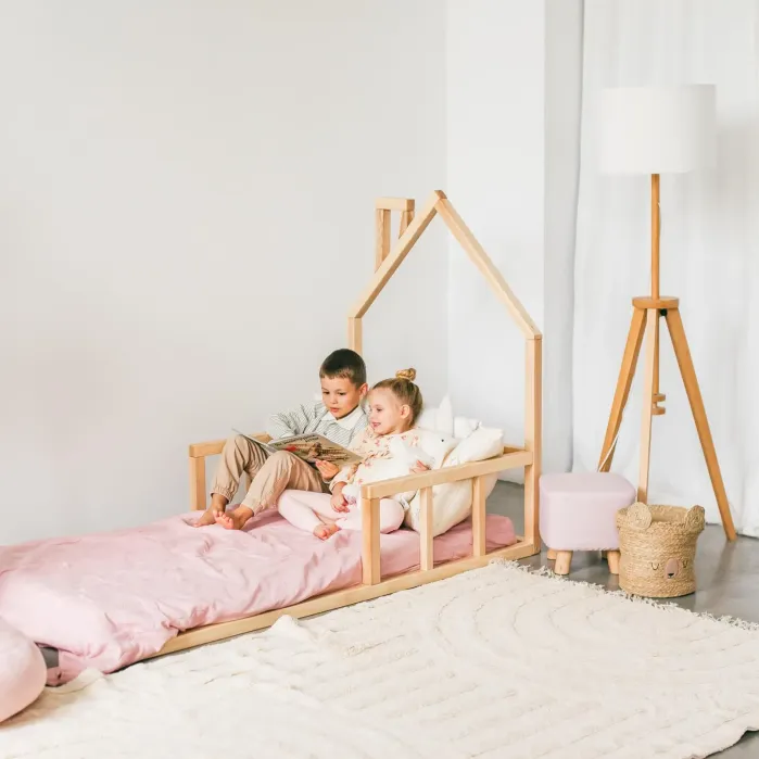 Two children sit on a wooden house-frame bed with pink bedding, reading a book together in a bright, minimalist room with a floor lamp and woven basket.