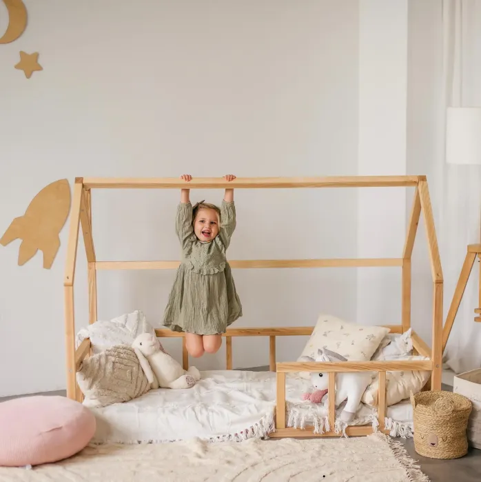 A young child in a green dress hangs from a wooden house-frame bed in a cozy room with plush toys, pillows, and soft decor.