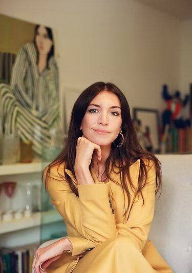 A woman with long brown hair, wearing a yellow outfit, sits indoors with her hand resting on her chin. A portrait of a woman in striped clothing is in the background.