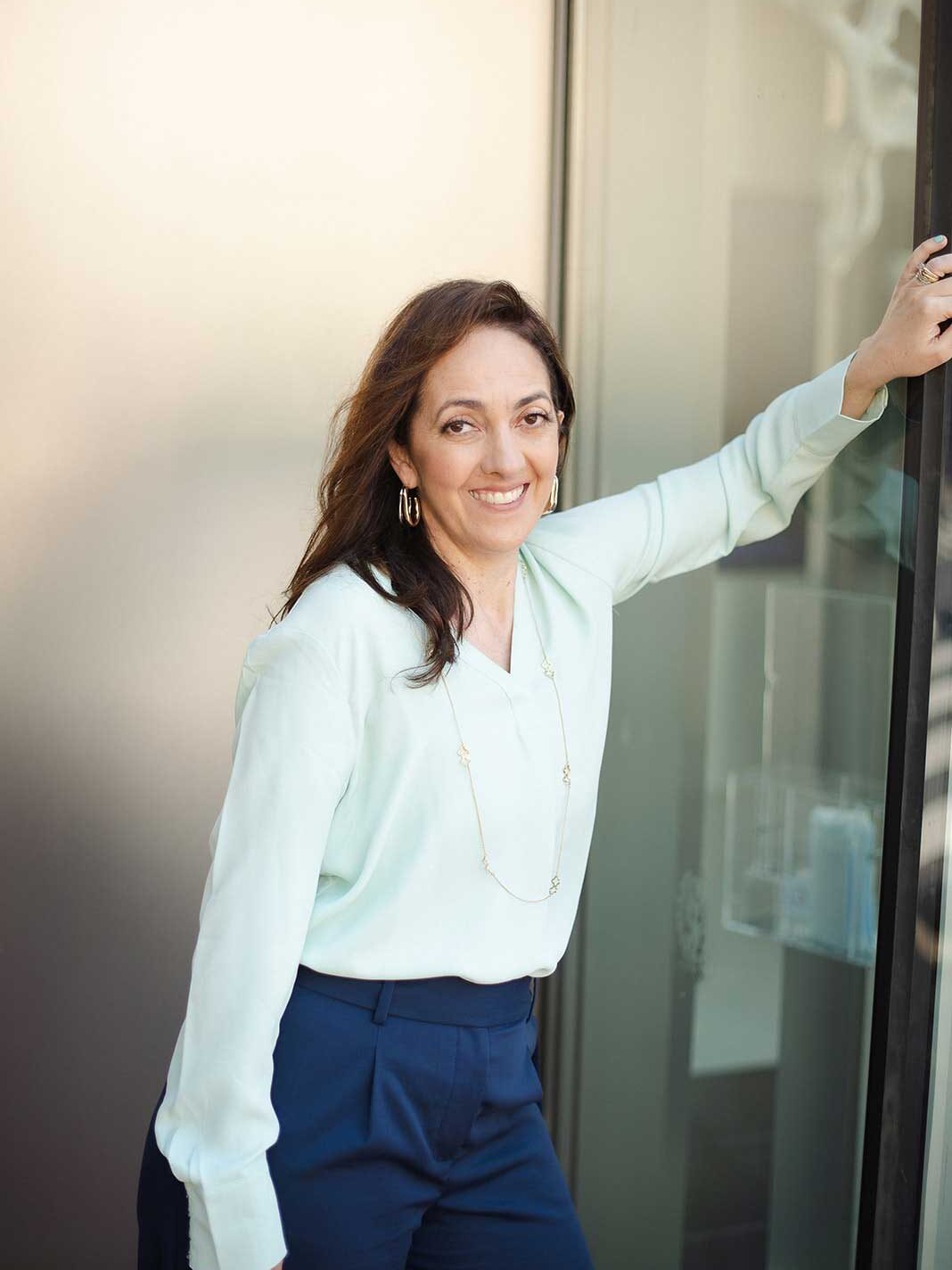 A woman with long brown hair, wearing a light blouse and blue pants, stands indoors leaning against a glass door, smiling at the camera.