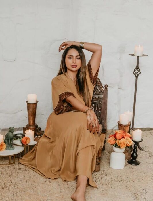 Woman in a tan dress sits barefoot on a wooden chair surrounded by candles, orange roses, and decorative items against a light-colored wall.