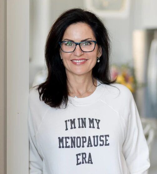 Woman with dark hair and glasses smiling, wearing a white sweatshirt that says "I'M IN MY MENOPAUSE ERA" in bold letters.