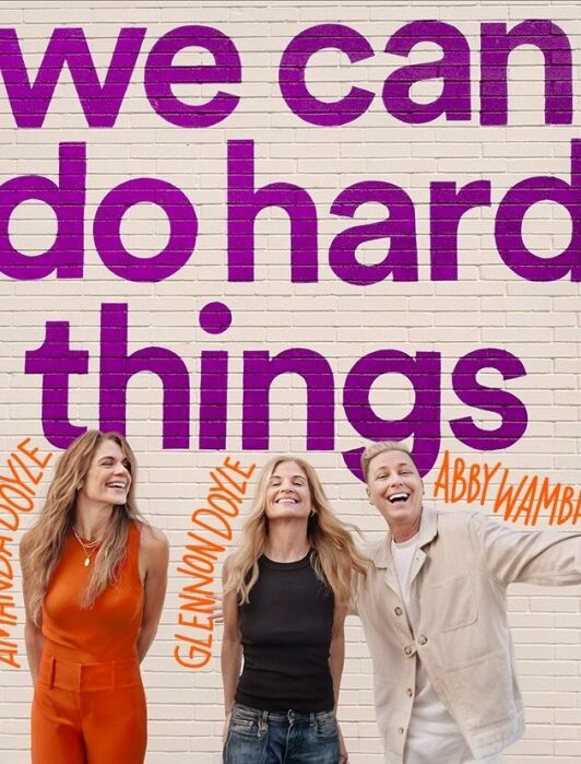Three women stand smiling in front of a brick wall with large purple text reading "we can do hard things"; their names, Amanda Doyle, Glennon Doyle, and Abby Wambach, are labeled in orange.