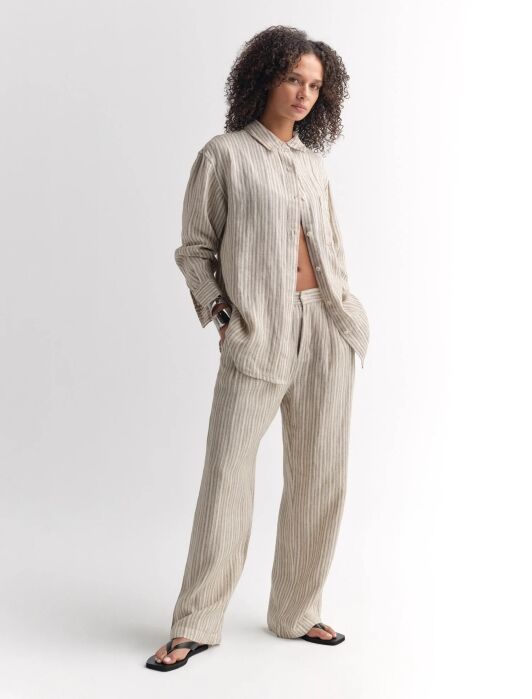 A person stands wearing a loose-fitting, beige and gray striped button-up shirt and matching wide-leg pants with black sandals against a plain white background.