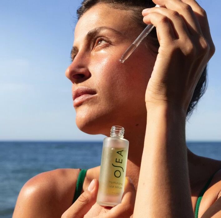 A woman applies serum to her face with a dropper while standing outdoors near the ocean; the serum bottle is labeled "OSEA Hyaluronic Sea Serum.
