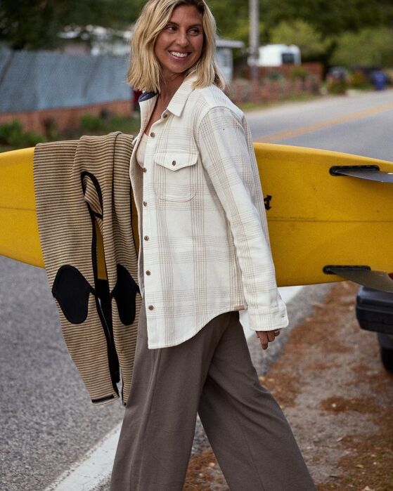 A woman stands outdoors next to a yellow surfboard, wearing a light plaid shirt and wide-leg pants, holding a striped towel.