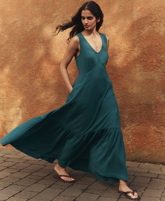 Woman in a sleeveless teal maxi dress and flip-flops walks on a paved path beside a textured, orange-brown wall.