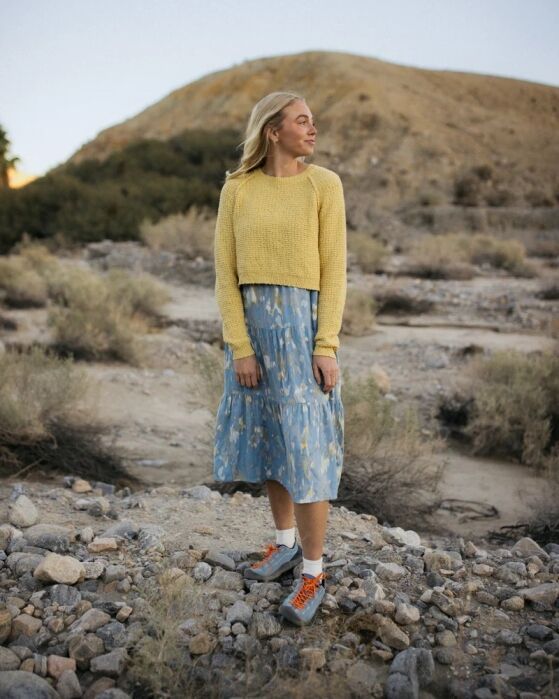 A woman stands on rocky ground outdoors, wearing a yellow sweater, a blue patterned skirt, and grey sneakers with orange laces. A hill and sparse vegetation are in the background.