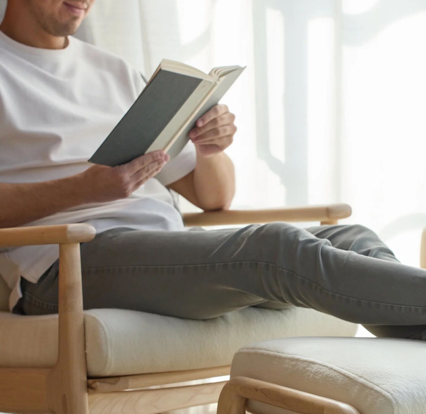 A person sitting in a wooden chair, wearing a white t-shirt and gray pants, reads a book in a brightly lit room.
