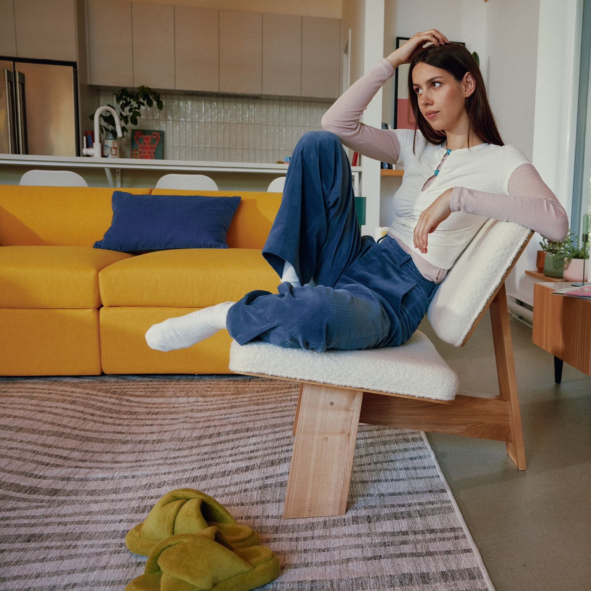 A woman sits on a modern lounge chair in a living room with a yellow sofa, striped rug, and green slippers on the floor.