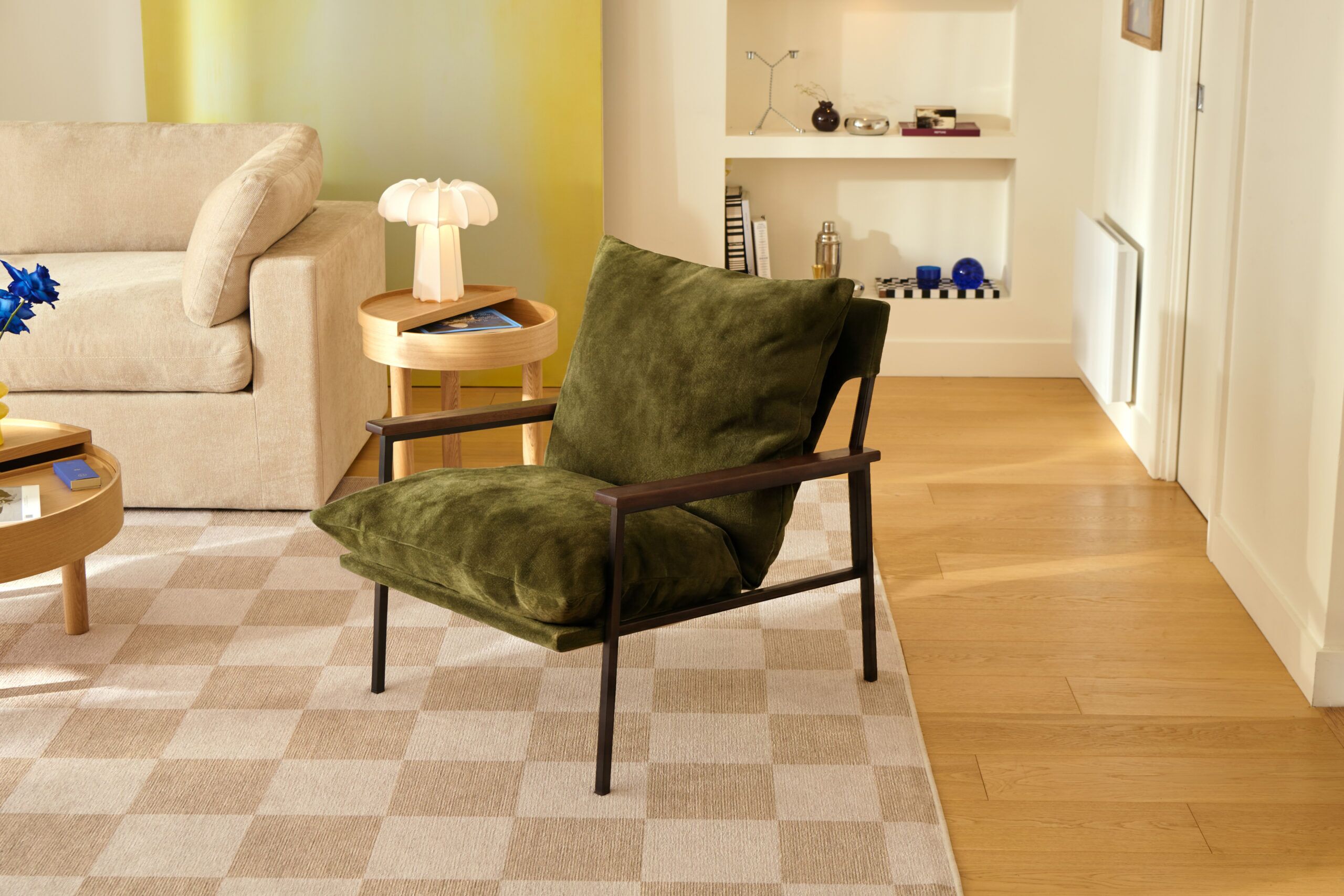 A green cushioned armchair with a black frame sits on a checkered rug in a modern living room with wooden floors and light-colored furniture.