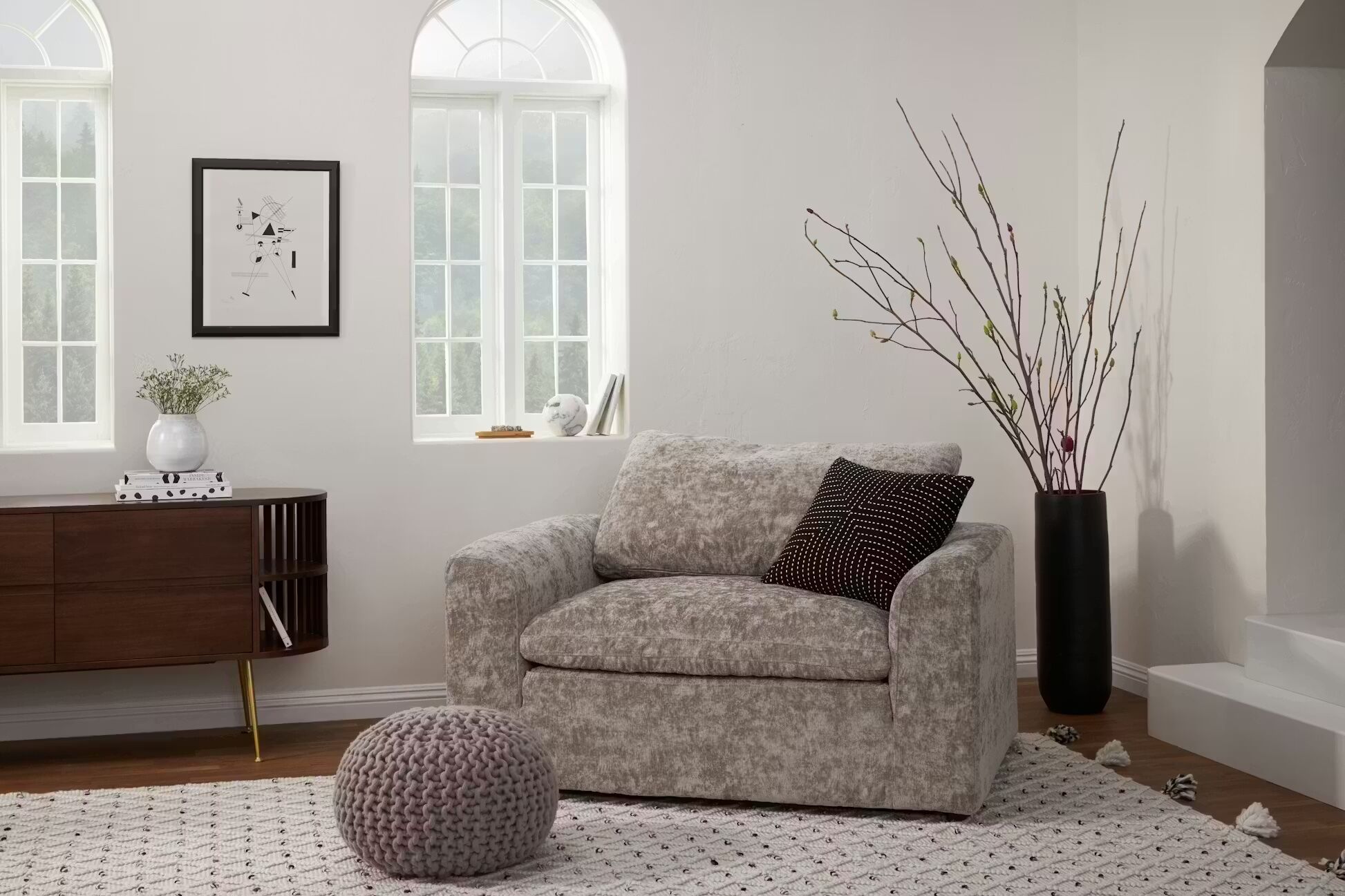 Modern living room with a gray armchair, black vase with branches, a wooden sideboard, a knitted pouf, and a patterned rug. Light enters through two arched windows.