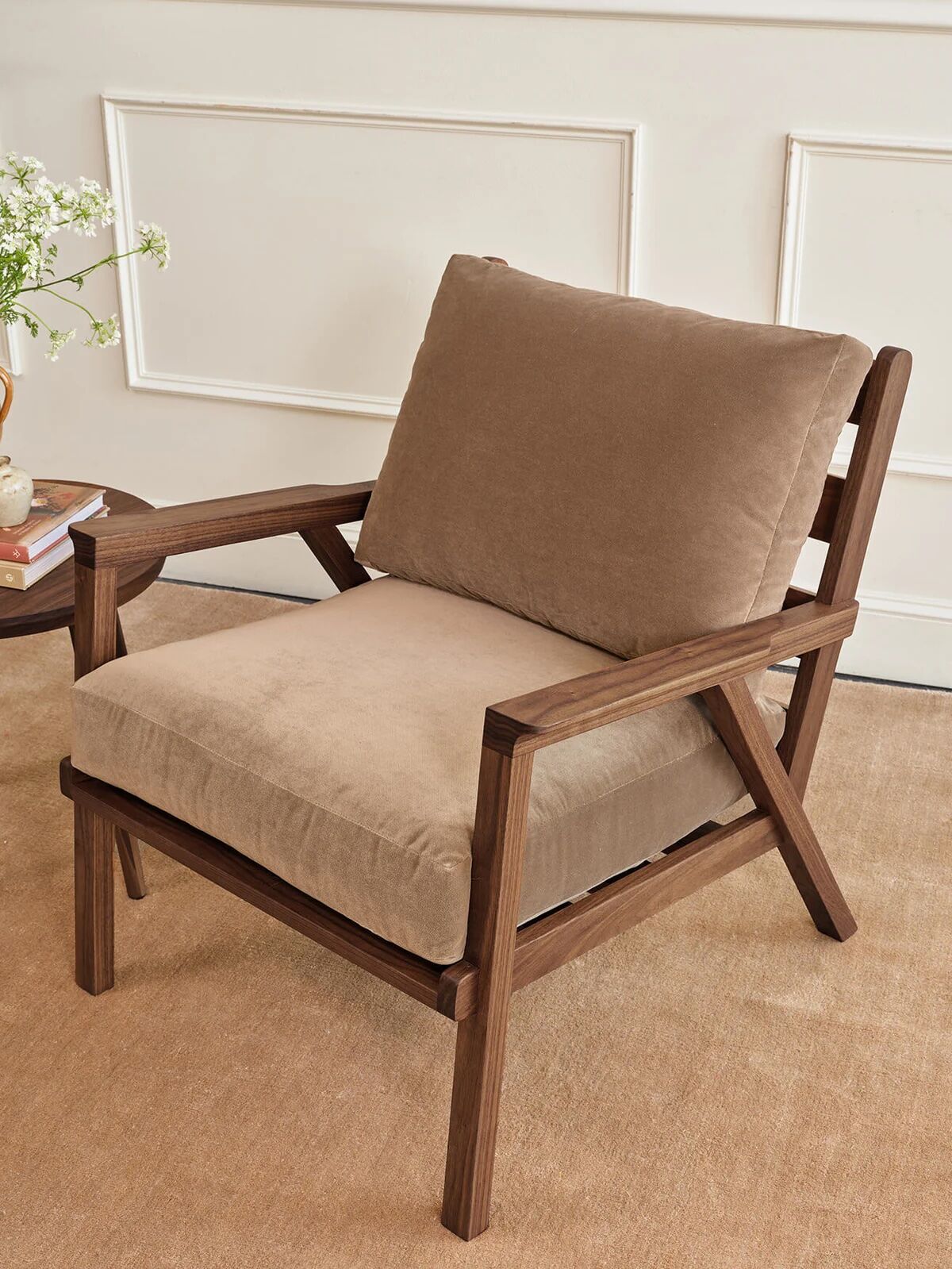 A cushioned armchair with a wooden frame and beige upholstery sits on a tan carpet next to a round side table with a vase of flowers.