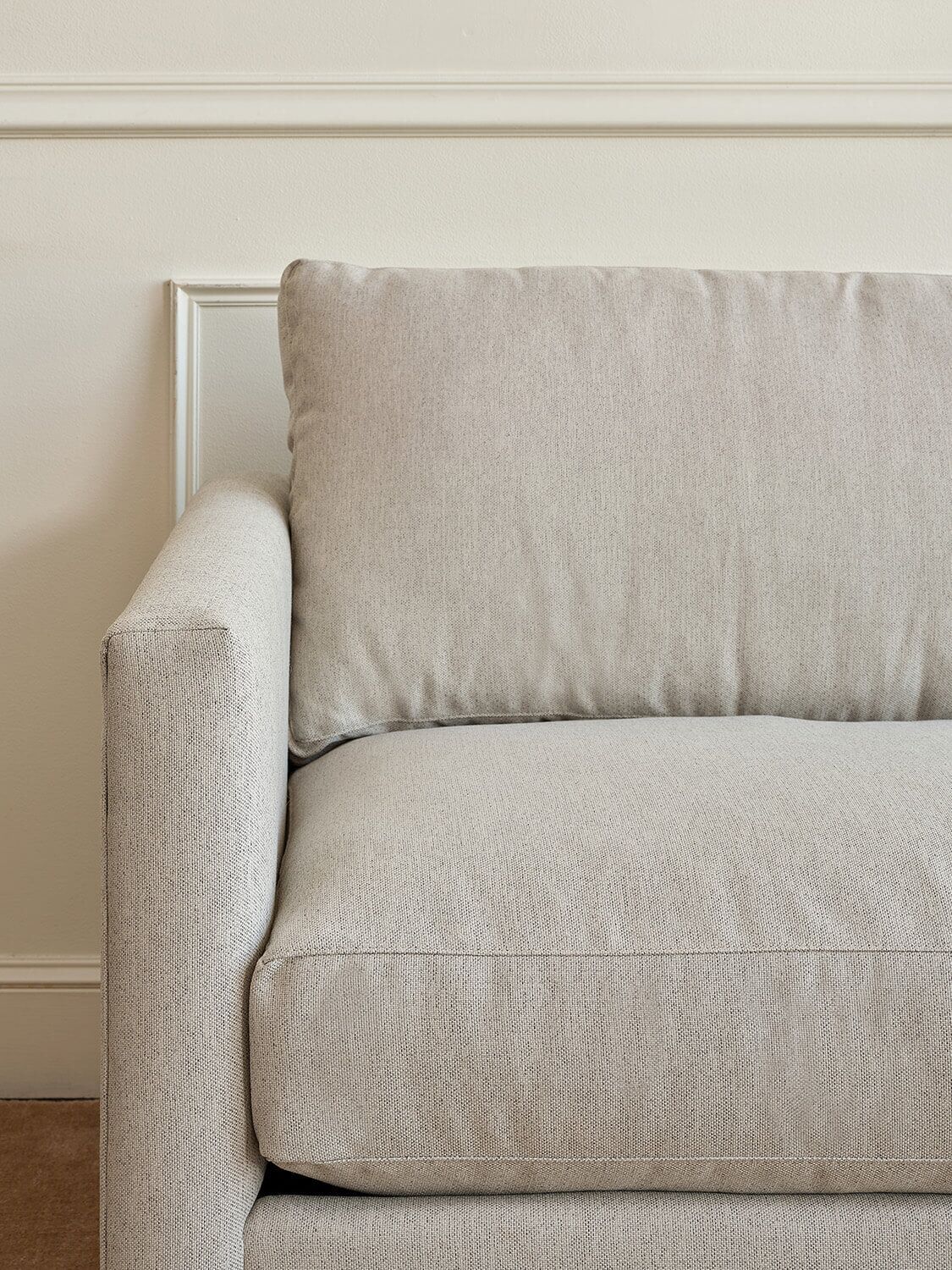 A close-up of a light beige modern sofa with a plush cushion, set against a cream-colored wall with decorative molding.