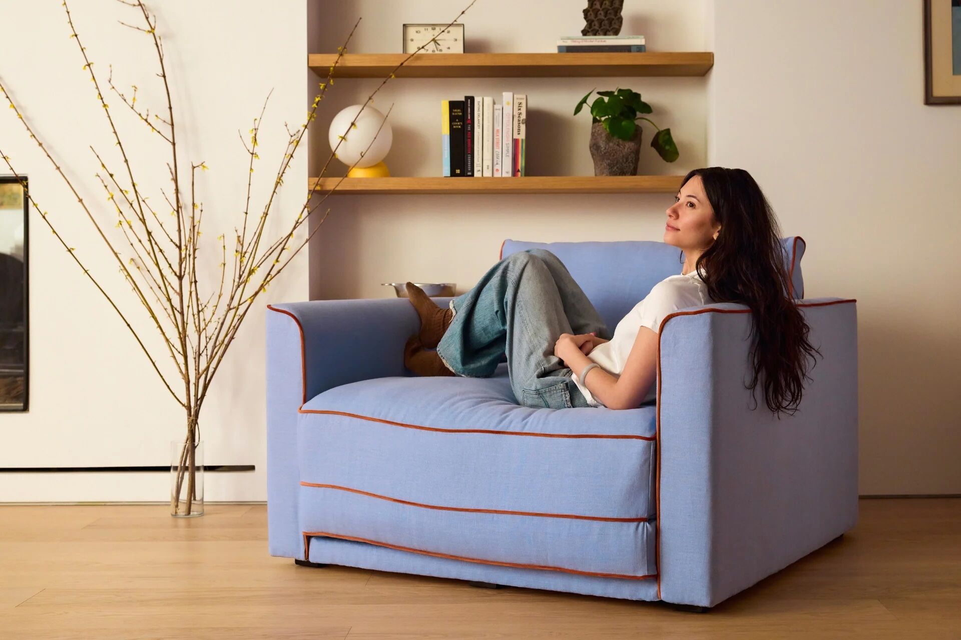 A woman in jeans and a white shirt sits relaxed on a large light blue armchair in a modern living room with shelves and decorative items in the background.