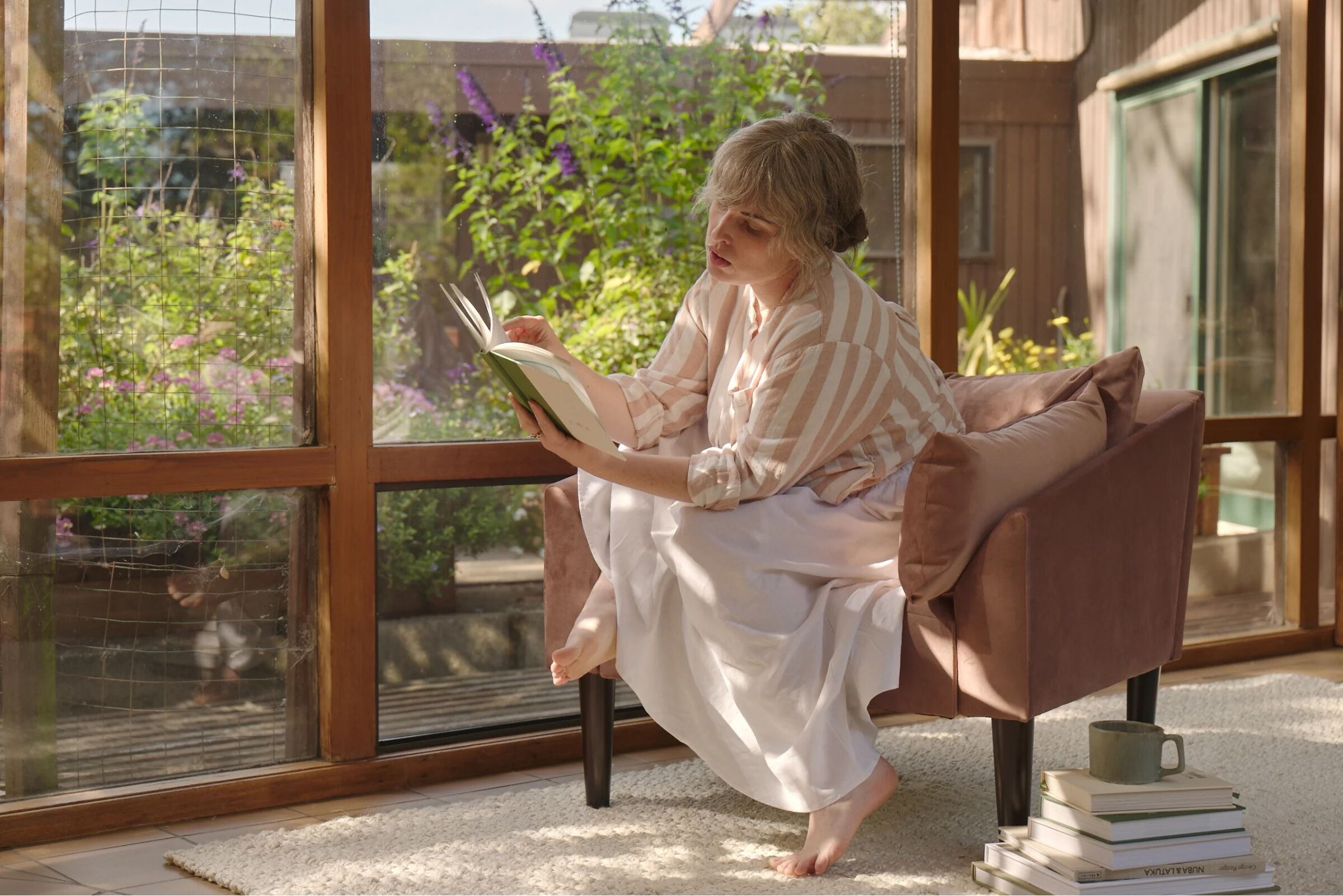 An older woman sits barefoot in a chair by large windows, reading a book. Sunlight streams in, with a garden visible outside. A mug and stacked books are on the rug nearby.