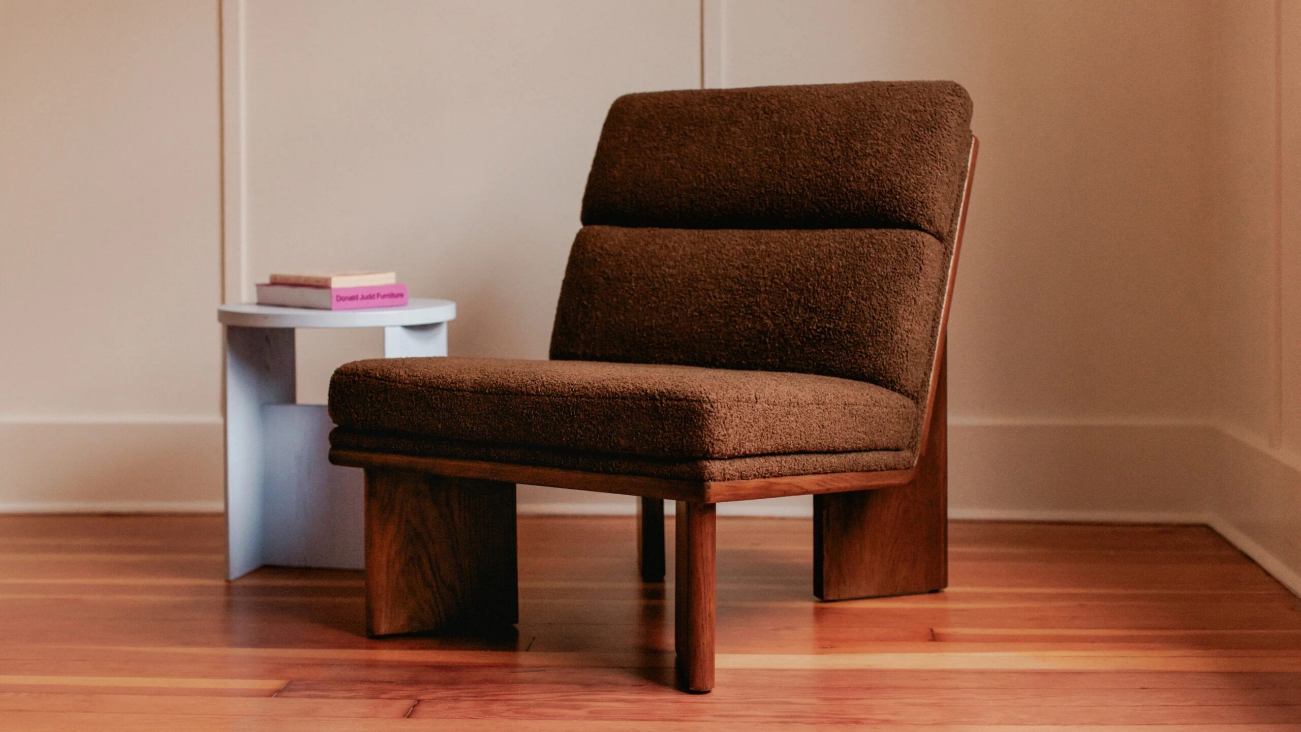 A brown cushioned lounge chair with wooden legs sits on a wooden floor next to a small white side table with two books.