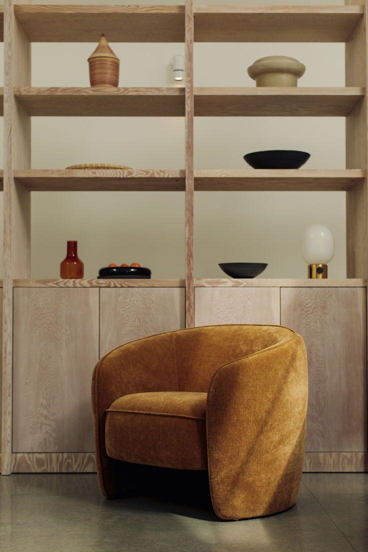 A mustard-yellow velvet armchair sits in front of light wood shelves displaying various decorative bowls, vases, and a lamp.