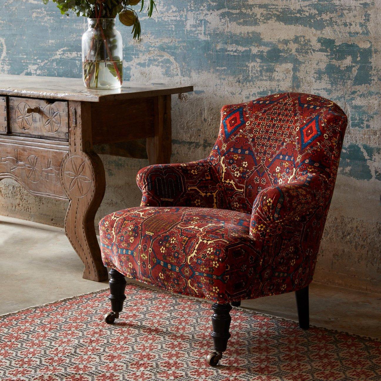 A red-patterned upholstered armchair sits on a matching rug beside a rustic wooden table with a glass vase of flowers, against a weathered blue wall.