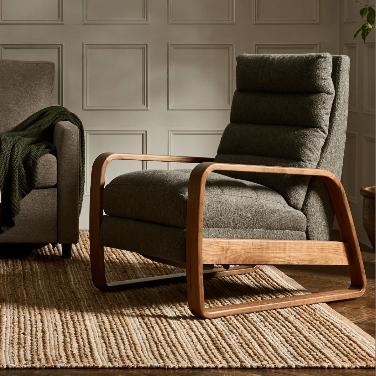 A modern gray cushioned lounge chair with wooden arms sits on a woven rug in a room with paneled walls and natural light.