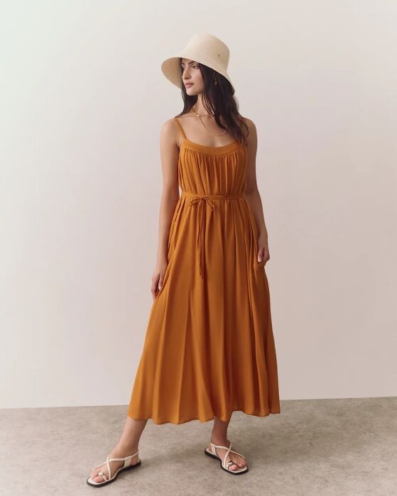 A woman in a sleeveless, mustard yellow maxi dress, white sandals, and a light-colored sun hat stands on a light gray floor against a plain background.