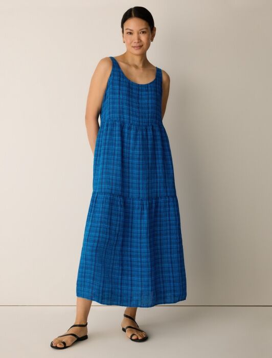 A woman stands against a plain background wearing a sleeveless, ankle-length blue plaid dress and black sandals.