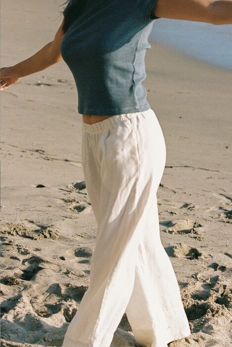 Person in a short-sleeve striped shirt and white pants walking on a sandy beach, with only their torso and legs visible.