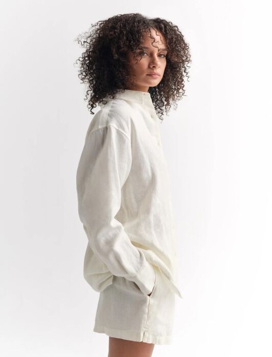 A person with curly hair wears a loose, long-sleeved, off-white button-up shirt and matching shorts, standing against a plain white background.