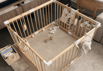 A wooden baby playpen with toys, balls, and a stuffed animal inside, set on a beige mat in a living room with books and a rocking chair nearby.