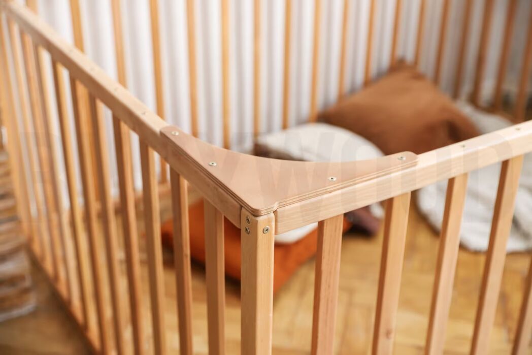 A wooden playpen with vertical slats, placed on a wooden floor, contains brown and white pillows inside.