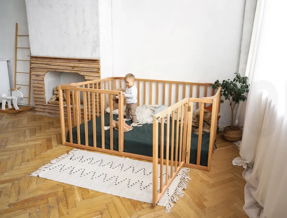 A small child stands inside a wooden playpen on a green mat, holding onto the bars in a sunlit room with wooden floors and minimalist decor.