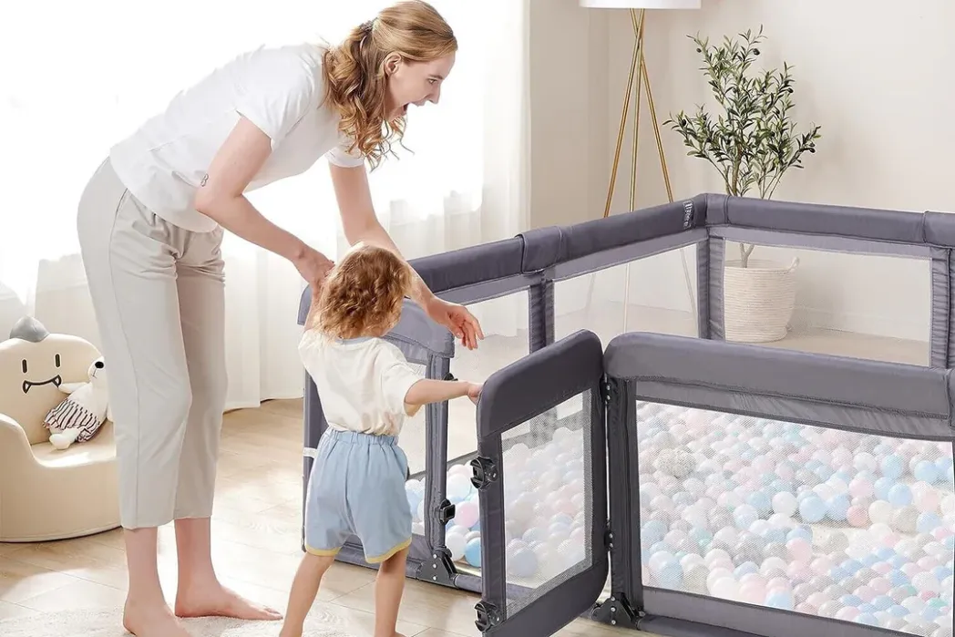 An adult helps a young child enter a mesh playpen filled with plastic balls in a bright, tidy room with a plant and a plush chair in the background.