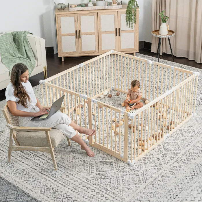 A woman works on a laptop while sitting near a playpen, where a baby plays with toys on a patterned rug in a living room.