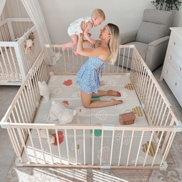 A woman kneels in a playpen, holding a baby up and smiling. The playpen contains stuffed animals and toys, with a crib and armchair visible in the background.