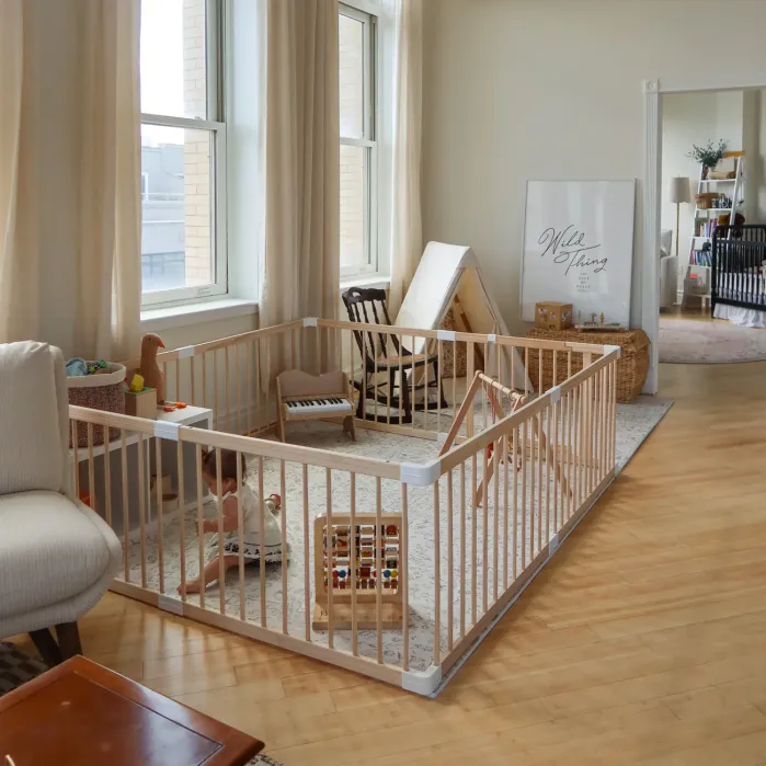 A playpen with wooden bars is set up in a bright, spacious room with large windows. Inside are toys, a baby, and a small rocking chair. The room has light wood floors and neutral decor.