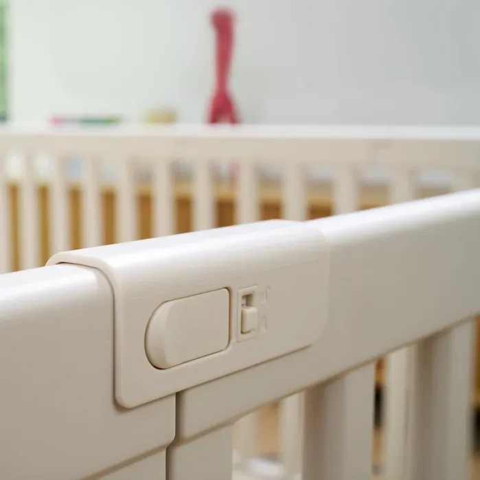 Close-up of a white plastic safety latch on the side of a baby crib, with blurred background showing crib bars and a red object.