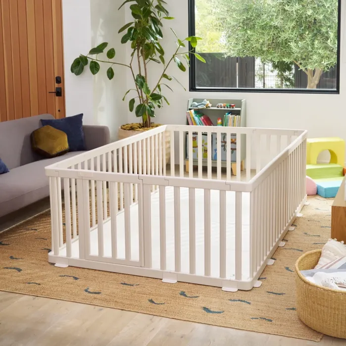 A white wooden playpen is set up on a woven rug in a living room with a sofa, indoor plants, and children’s toys and books in the background.