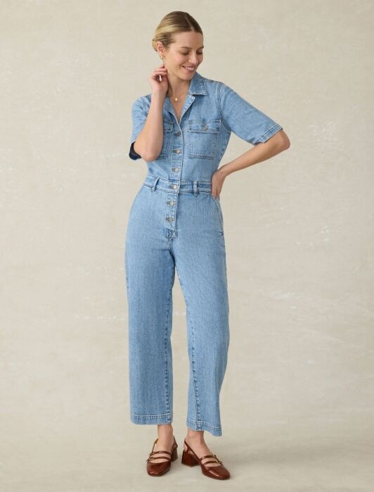 A woman wears a short-sleeved light wash denim jumpsuit and brown Mary Jane shoes.