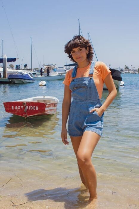 A person wearing an orange t-shirt and denim overall shorts stands in the shallow water of a marina, with boats in the background.