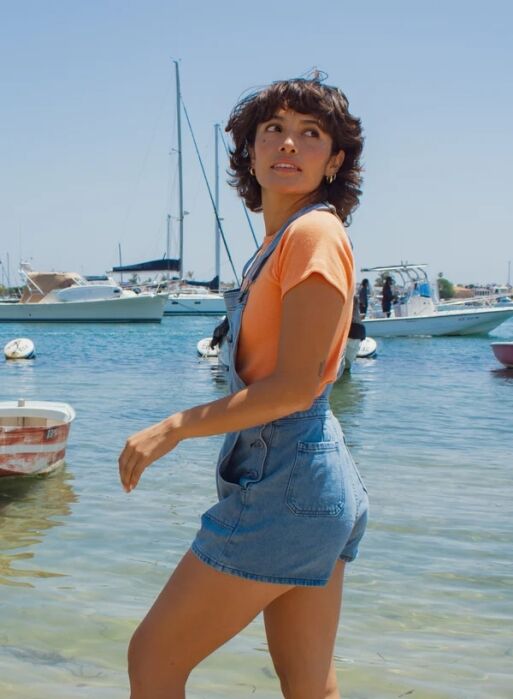 A person wearing a peach t-shirt and denim overall shorts stands in front of a harbor with sailboats.