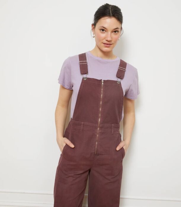 A person wearing a light purple t-shirt under brown zippered overalls with their hands in their pockets.