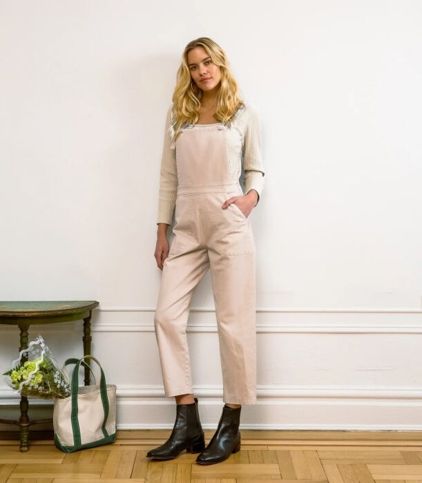 A woman wearing beige overalls over a cream long-sleeved shirt and black ankle boots.