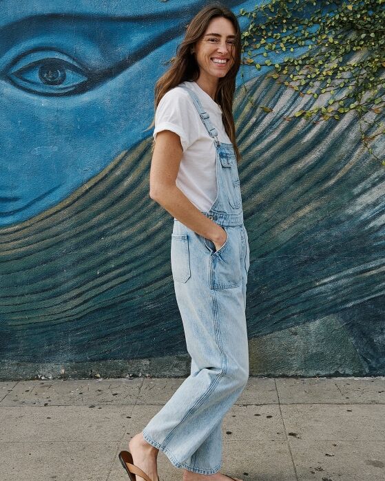 A smiling woman wearing a white t-shirt and light-wash denim overalls stands in front of a blue mural.